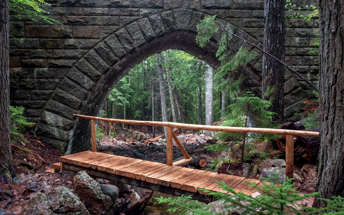 A newly constructed wooden bridge crosses a river in a forest with a stone arched bridge behind it.