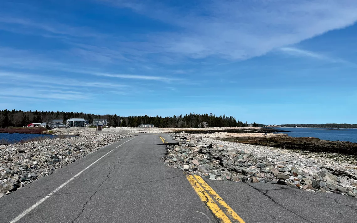 One lane of a flat section of asphalt road along the seashore is torn apart, with rocks where the asphalt was. There are some buildings in the background.