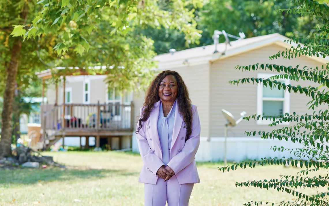 Catherine Coleman Flowers wears a lavender pantsuit and stands in front of a house, smiling at the camera.