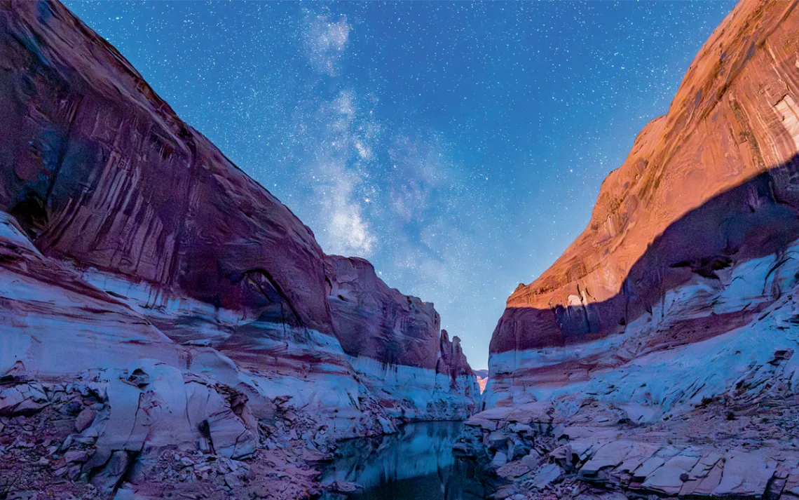 Early evening photo of a canyon in a low Lake Powell, with a white layer at the bottom and a redrock layer up top. A small boat is beached on the shore.