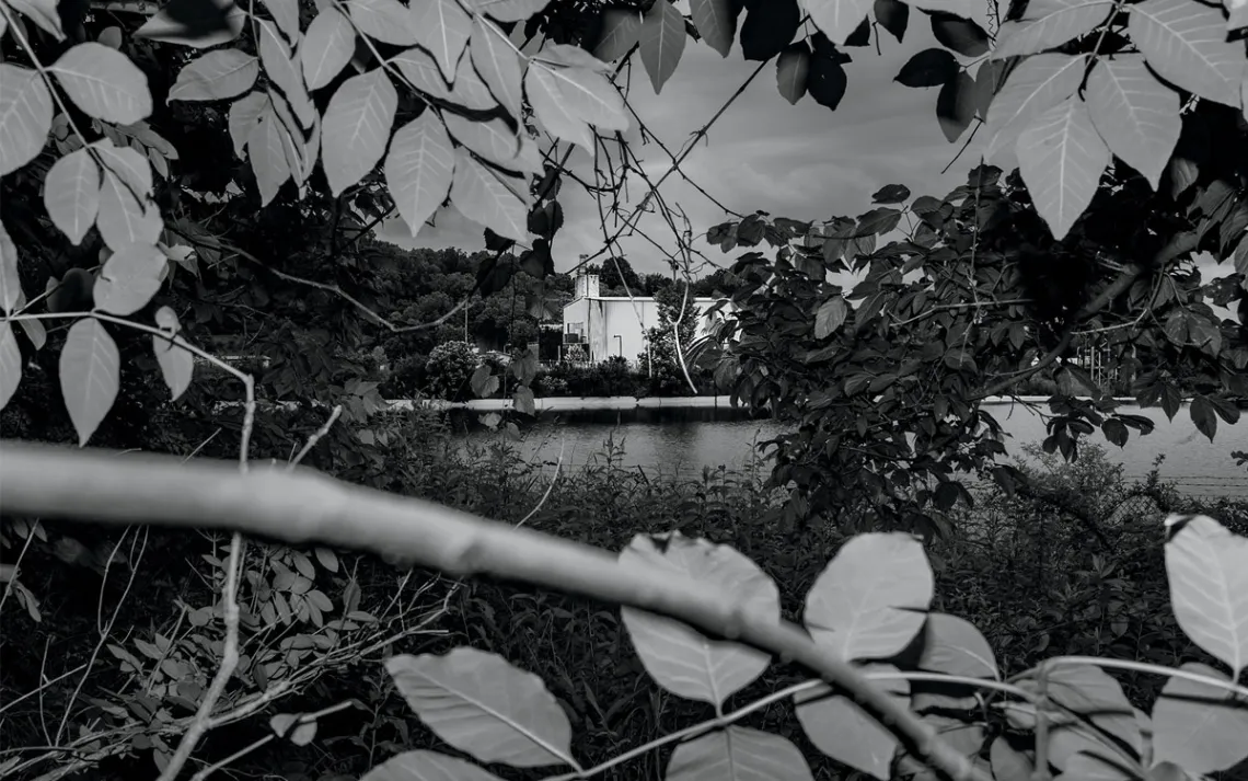  A black-and-white photo taken from bushes across a waterway from a white building.