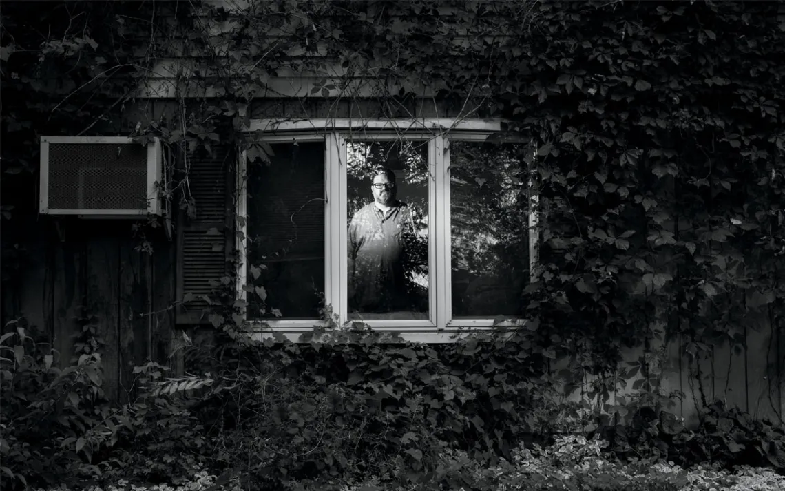  Black-and-white photo of Sean Guthrie looking at the camera through a window inside his house. The house has vines covering it.
