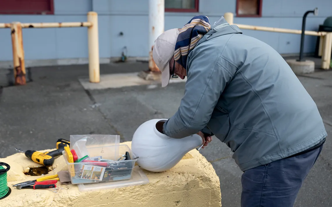 Carl Bevins wears cold-weather clothing and has his hand inside a white gourd. A box of tools sits next to it.