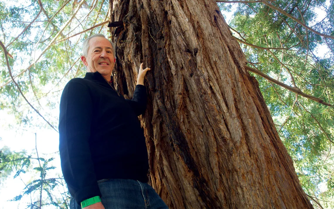 Rick Lanman stands with his left hand on a redwood tree trunk as he looks down at the camera.