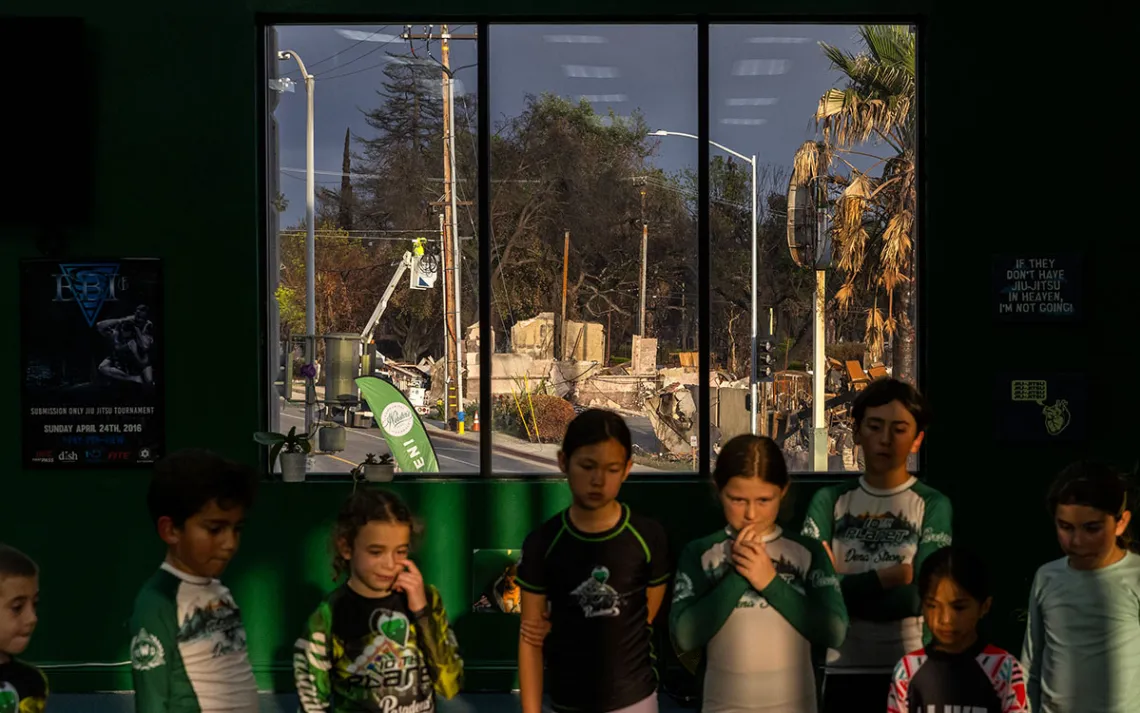 Children stand in the foreground at a jiujitsu class. In the background out the window, you can see rubble from their burned neighborhood.
