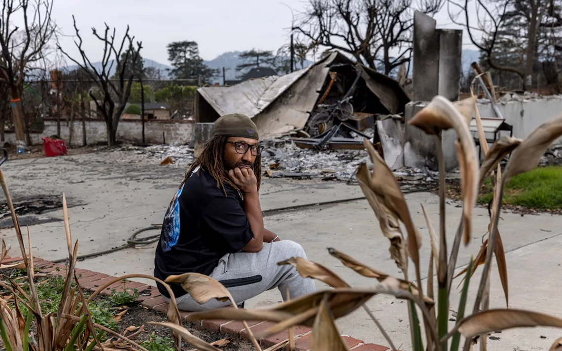 A man sits facing the camera, among the burned rubble of his home