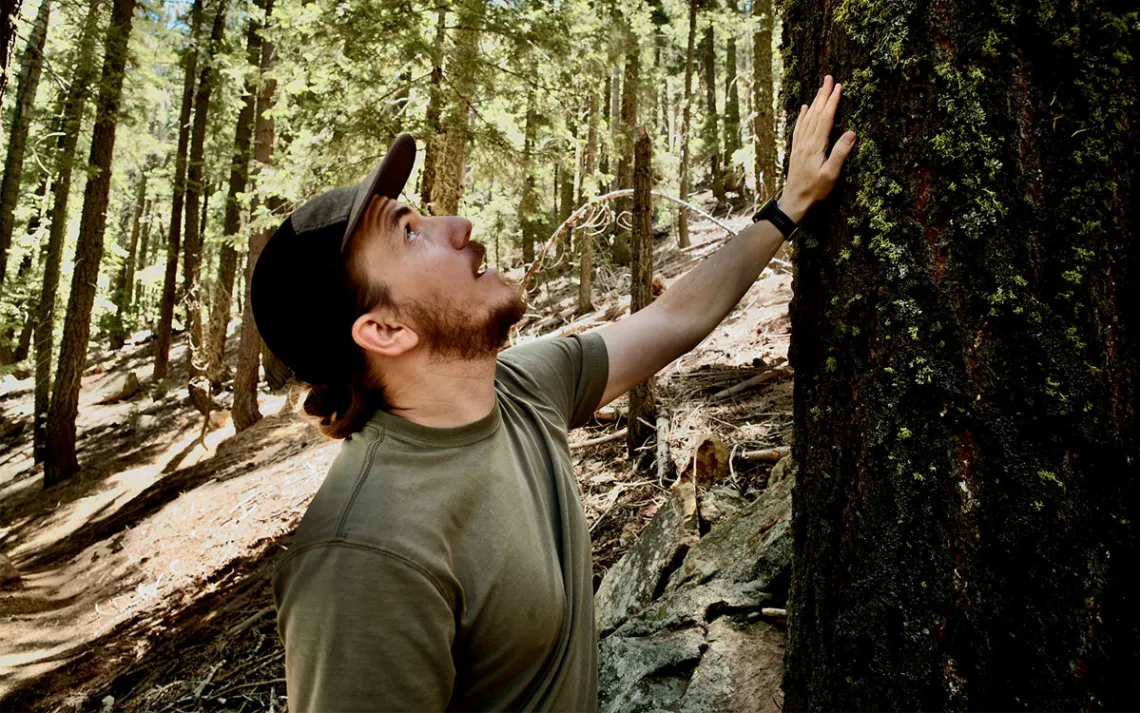 Side view of Bryant Baker, wearing a dark-green hat and shirt, placing his hand on a tree in a wooded area of Los Padres National Park.