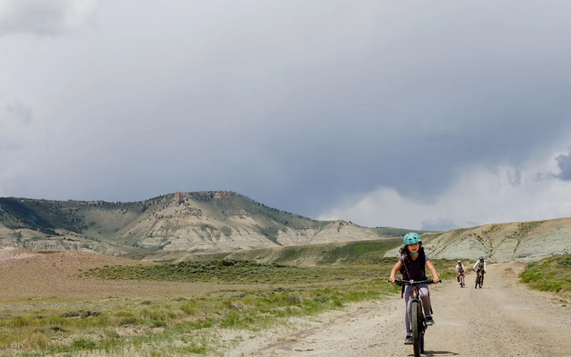 Miriam Peterson wears a turquoise helmet and rides on an unpaved road toward the camera with two other riders and the gray, rocky Oregon Buttes behind her.