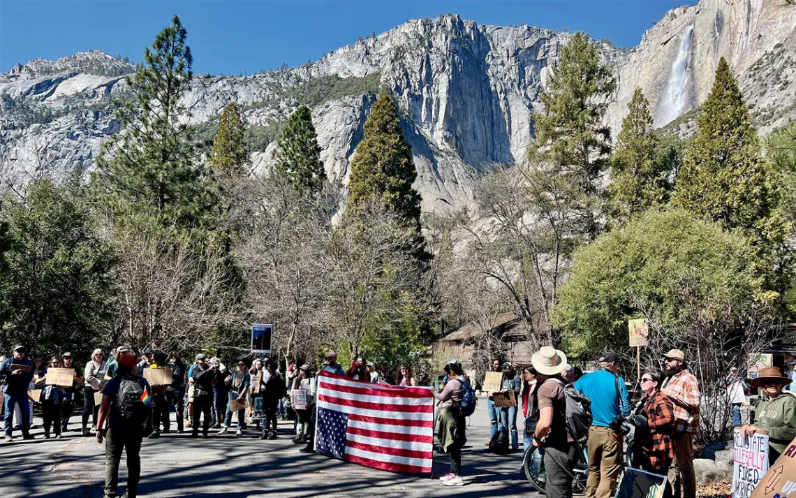A line of protesters hold signs and an upside-down US flag in Yosemite National Park in front of a waterfall.