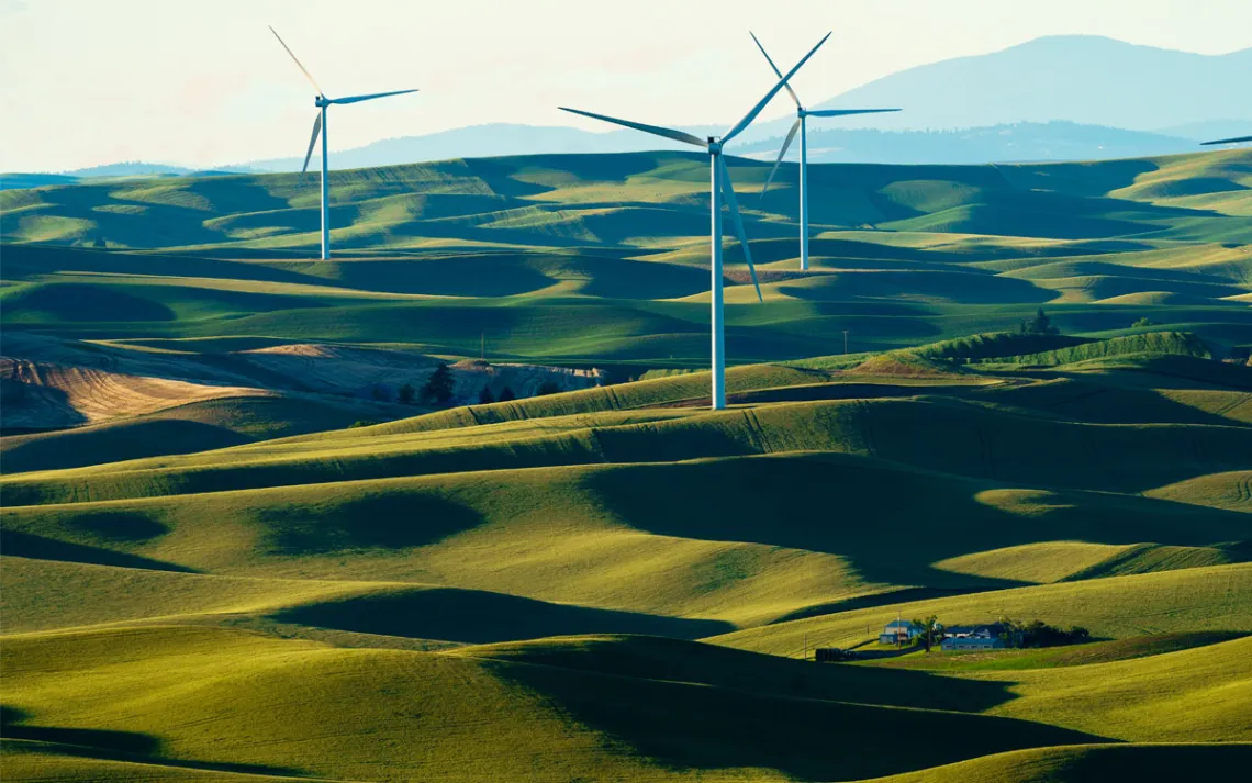 Three windmills stand to attention in a lush, rolling green field, with hills and mountains in the background