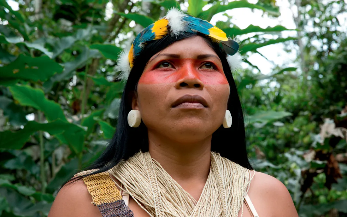 Close-up of Nemonte Nenquimo looking into the distance, wearing a feather headdress and string top. A red stripe is painted across her eyes and nose.