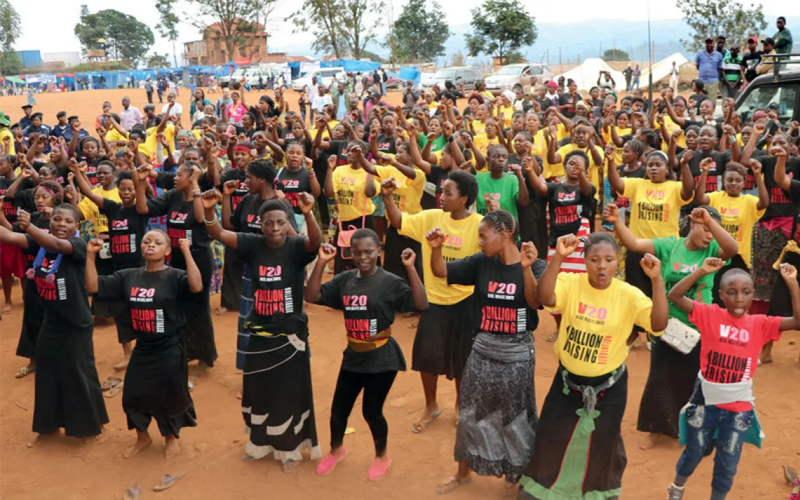 More than 100 Black women wearing different-colored "V20 1 Billion Rising" T-Shirts dance with their arms in the air on a reddish-dirt ground.