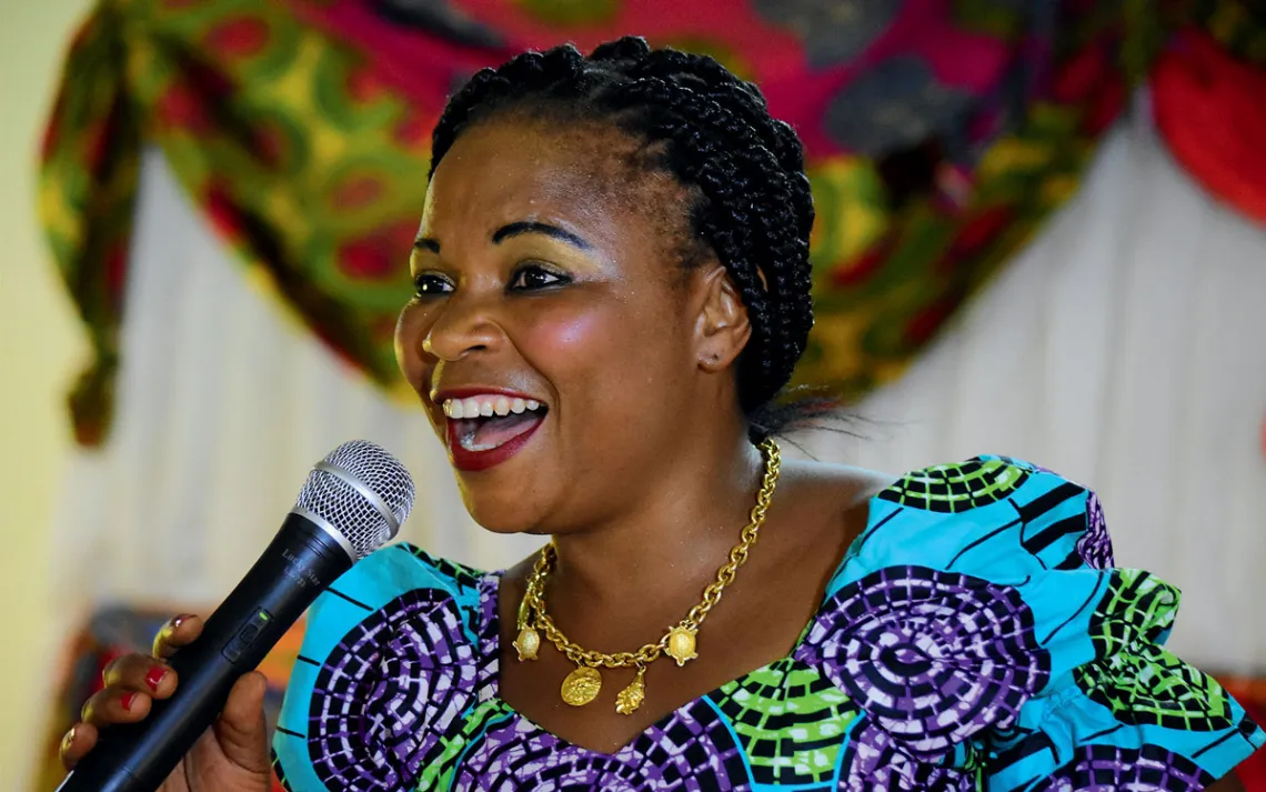  Jane Mukunilwa, wearing a bright dress and gold necklace, smiles and sings into a microphone.