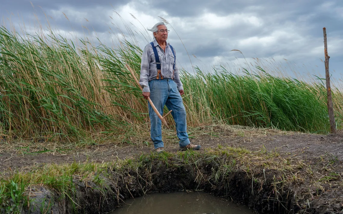 Jim Enote wears jeans and suspenders and stands atop a spring surrounded by tall grass. He's holding a long wooden instrument.