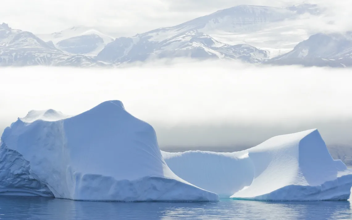 A large iceberg floats just off shore in Baffin Bay, Greenland. A bank of fog lies between the iceberg and the mountains in the background.