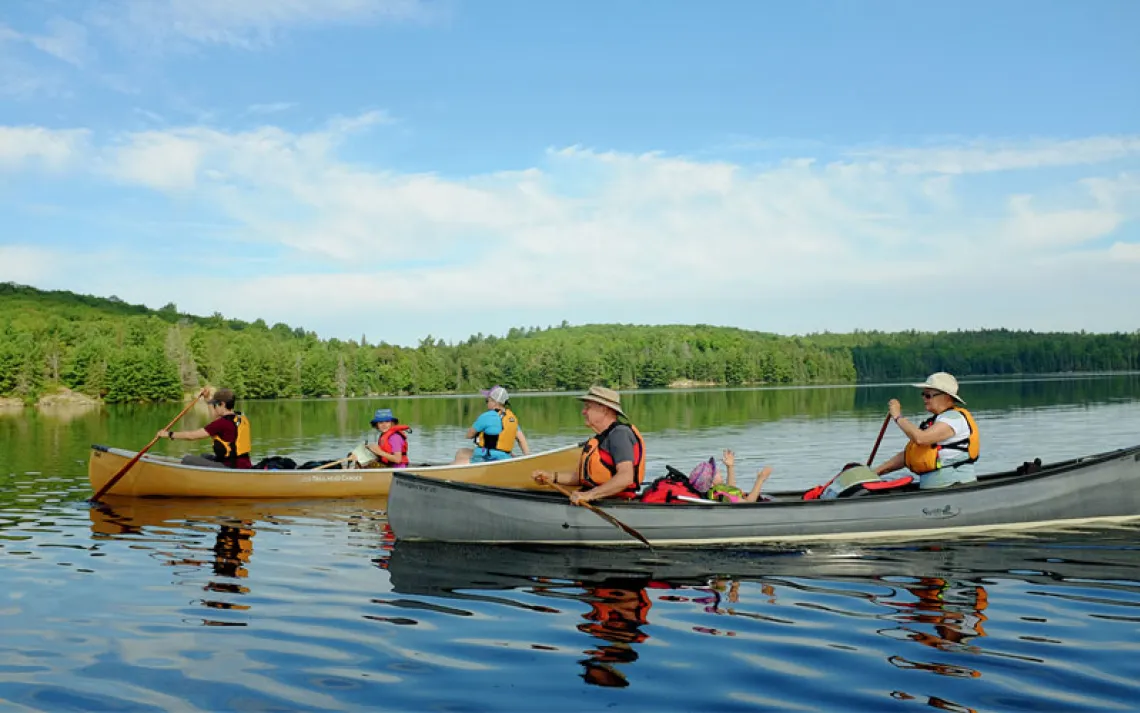 Four adults and two children travel in two canoes across a calm lake with trees in the background