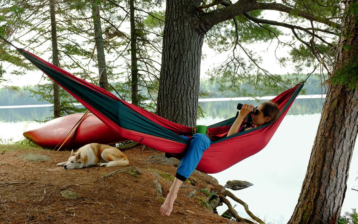 A woman lies in a red hammock next to a lake and looks out binoculars while a light-colored dog sits nearby