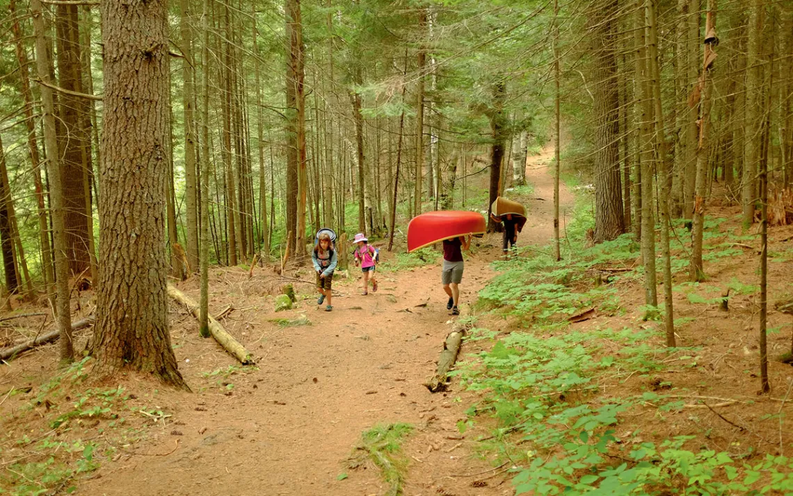 Two adults carry a red and gold canoe while two kids carry a backpack and food canister up a treelined trail