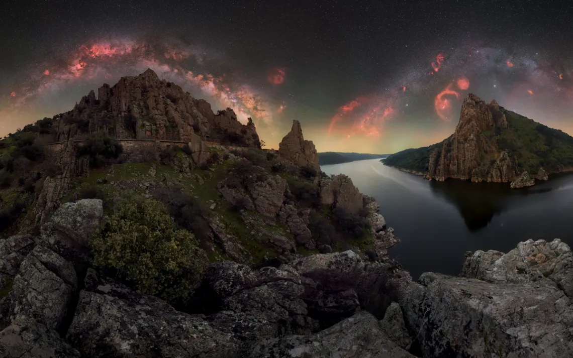 A river runs through rocky mountains with orange swirls of the Milky Way above in Spain's Monfragüe National Park.