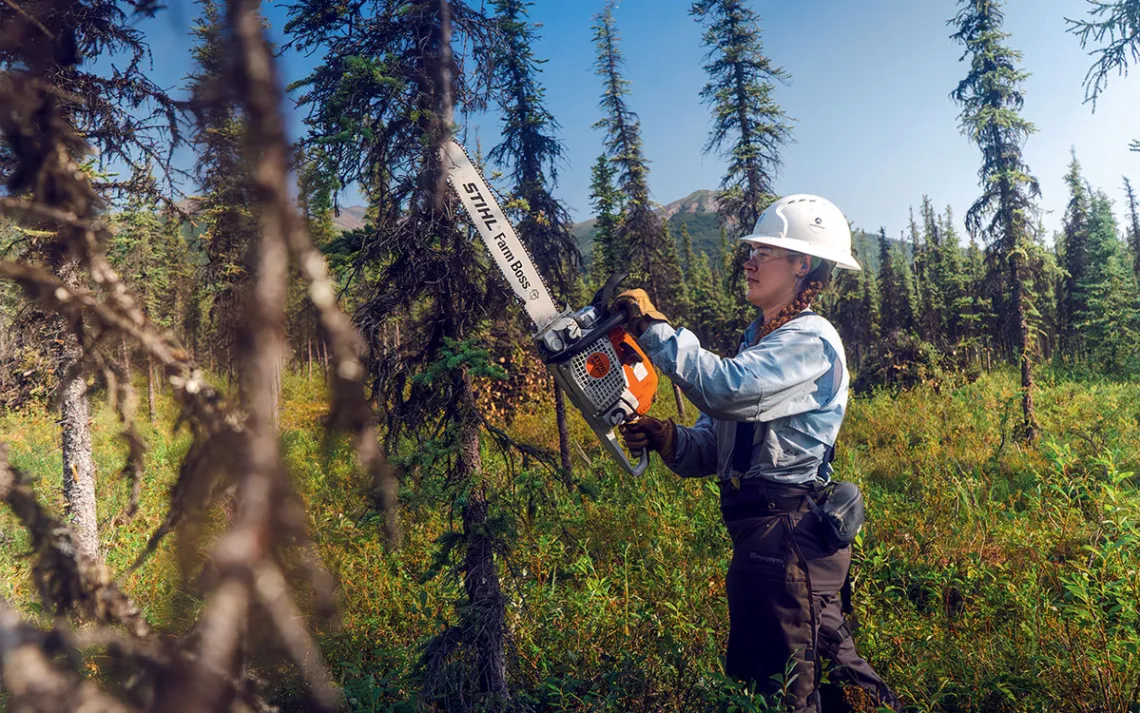 A woman wearing a white hard hat holds up a chainsaw next to a tree.