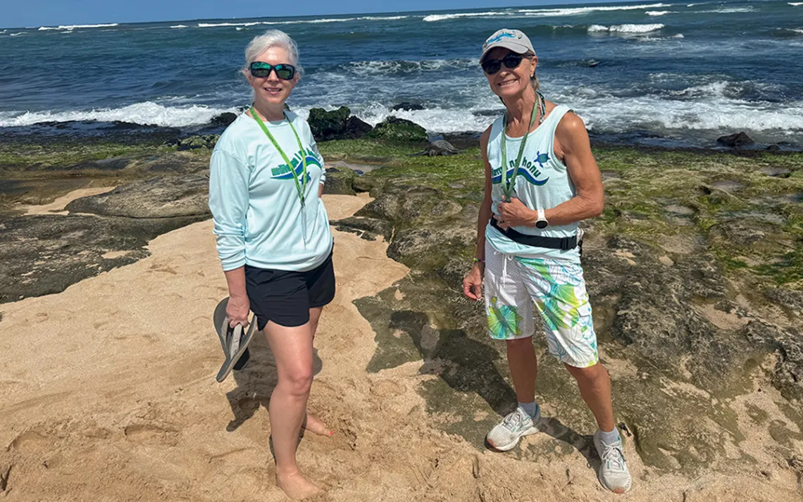 Two people stand on a volcanic-rocky beach with the ocean behind them