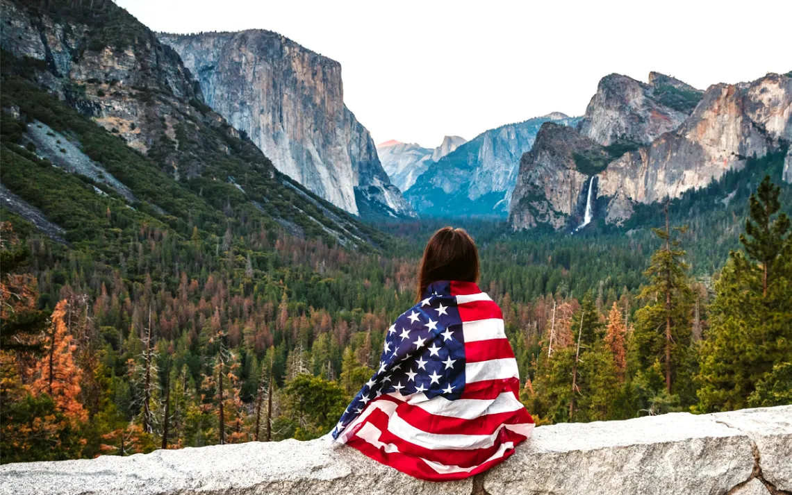 The back of a person draped in an American flag looking out at a viewpoint in Yosemite National Park, including a waterfall and Half Dome.