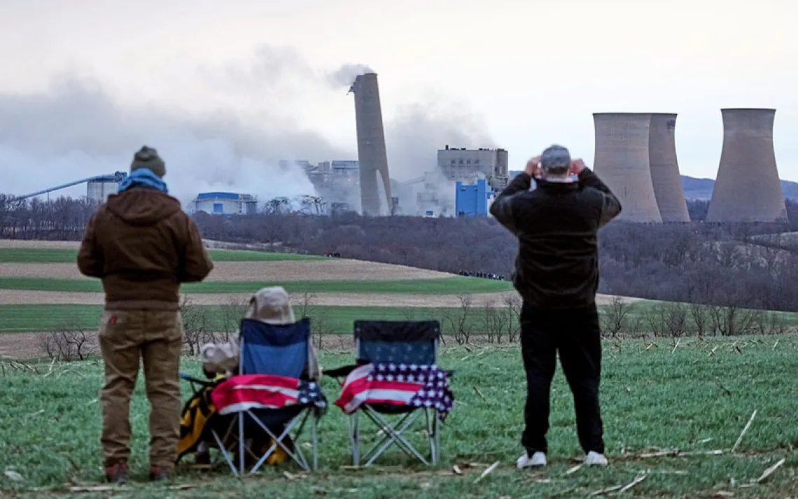The backs of three people with US flags on their chairs watching the demolition of the Homer City Generating Station in the distance.