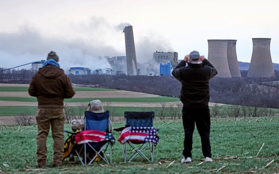 The backs of three people with US flags on their chairs watching the demolition of the Homer City Generating Station in the distance.