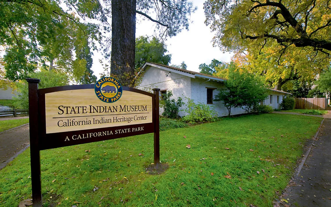  A sign reading "State Indian Museum California Indian Heritage Center / A California State Park" sits in front of a one-story painted-white adobe building with trees surrounding it.