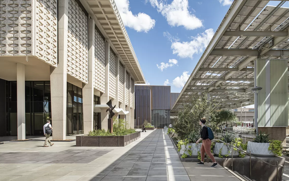 Students walk on a pathway next to a 1950/60s concrete building. To the right is a newly built outdoor area protected by panels that may be solar.