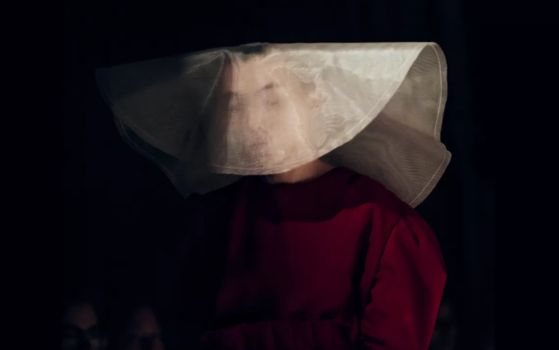 A woman wears a conservative burgundy dress and translucent white headdress inspired by The Handmaid's Tale.