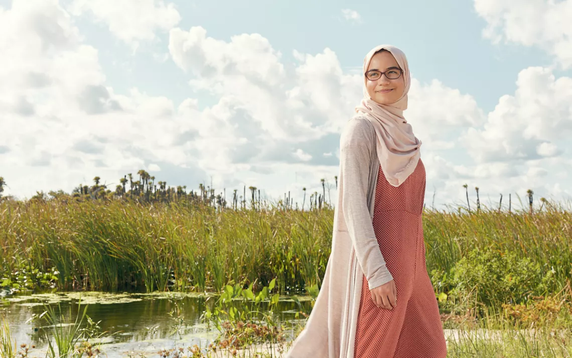 Tasnim Mellouli wears a cream hijab and cardigan and a rust-colored dress. She's wearing glasses and looking to her right, smiling. Behind her is a swampy landscape.