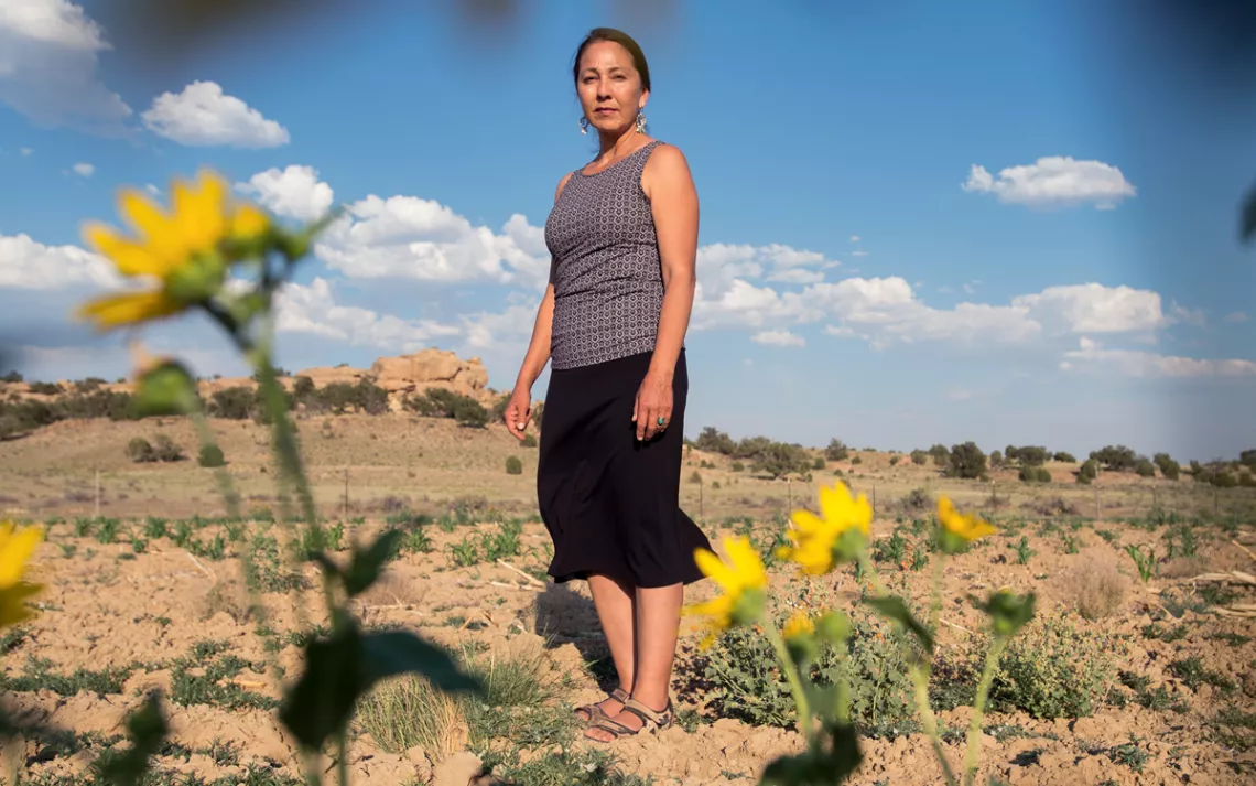Nicole Horseherder is wearing a skirt and tank top and standing on a desert landscape, looking at the camera.