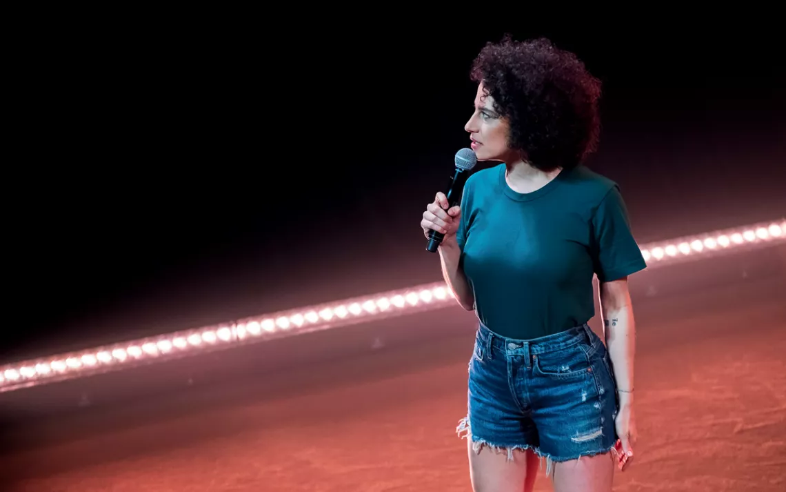 Ilana Glazer is on stage, holding a mic and looking to her right. She's wearing cut-off jean shorts and a teal T-shirt.