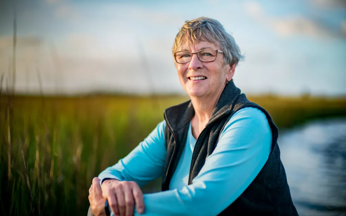 Penny Hooper sits with her arms crossed on her knee. She's wearing glasses, a blue shirt, and a black fleece vest.