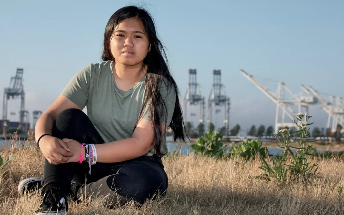 Angelika Soriano sits on dry grass with her arms around one knee, looking at the camera. In the background is the Port of Oakland.