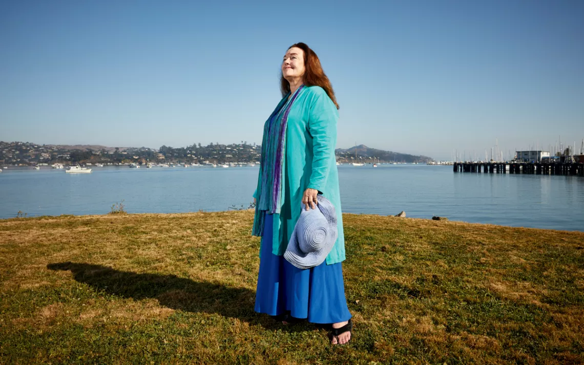 Mary Crowley, wearing bright blue and turquoise, stands by the bay in Sausalito.