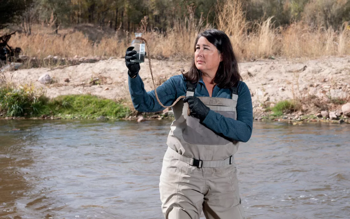 Fran Silva-Blayney stands in the middle of a river, wearing waders and holding up a water sample.
