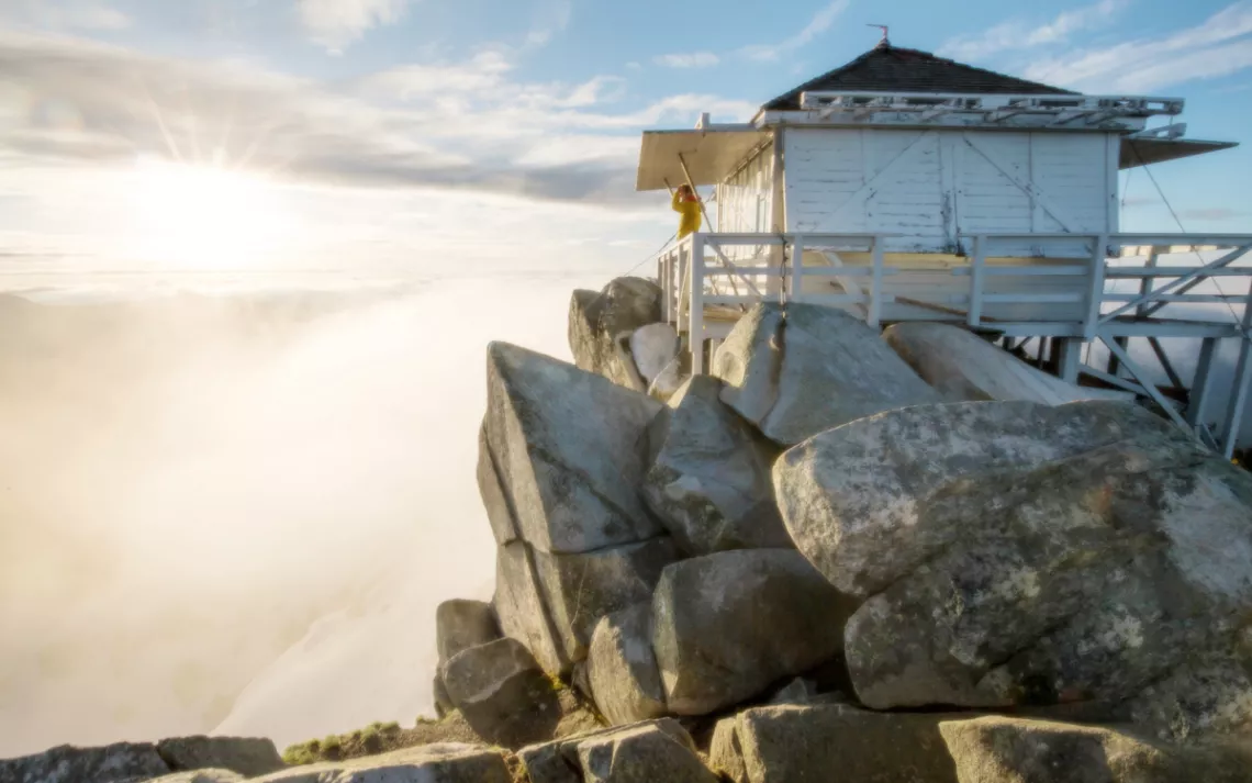 A hiker watches the sunrise from the porch of the Mt. Pilchuck fire lookout in Washington. The building is a white weathered cabin atop boulders.