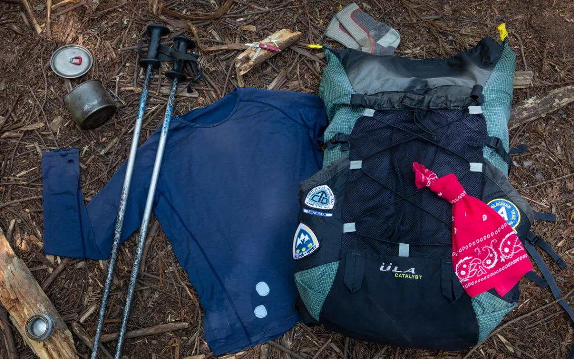 An ULA backpack and the author's accessories sit in front of a redwood tree in a forest location.