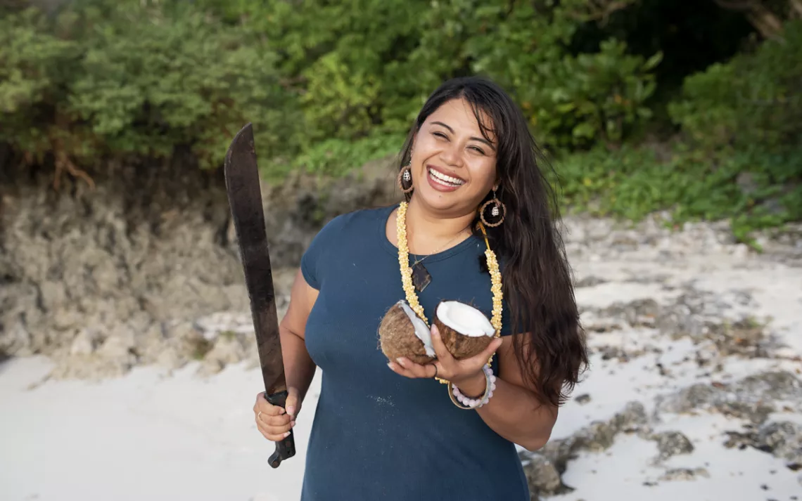 Moñeka De Oro smiles and stands on a beach holding a cut coconut and a machete.