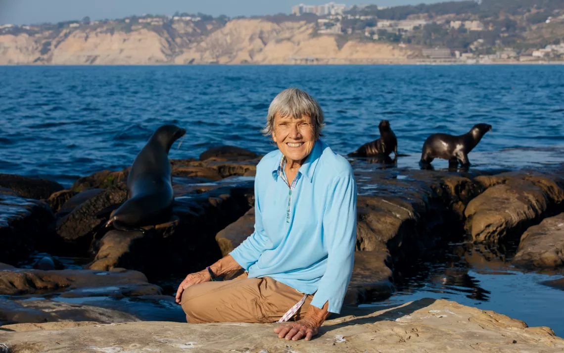 Ellen Shively sits on a rock by the ocean. Behind her are several seals.