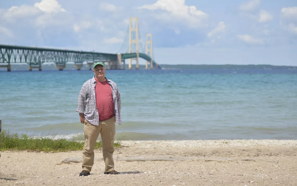 David Holtz stands on a beach with the Mackinac Bridge in the background. He's wearing a baseball cap, an unbuttoned shirt, and khakis.