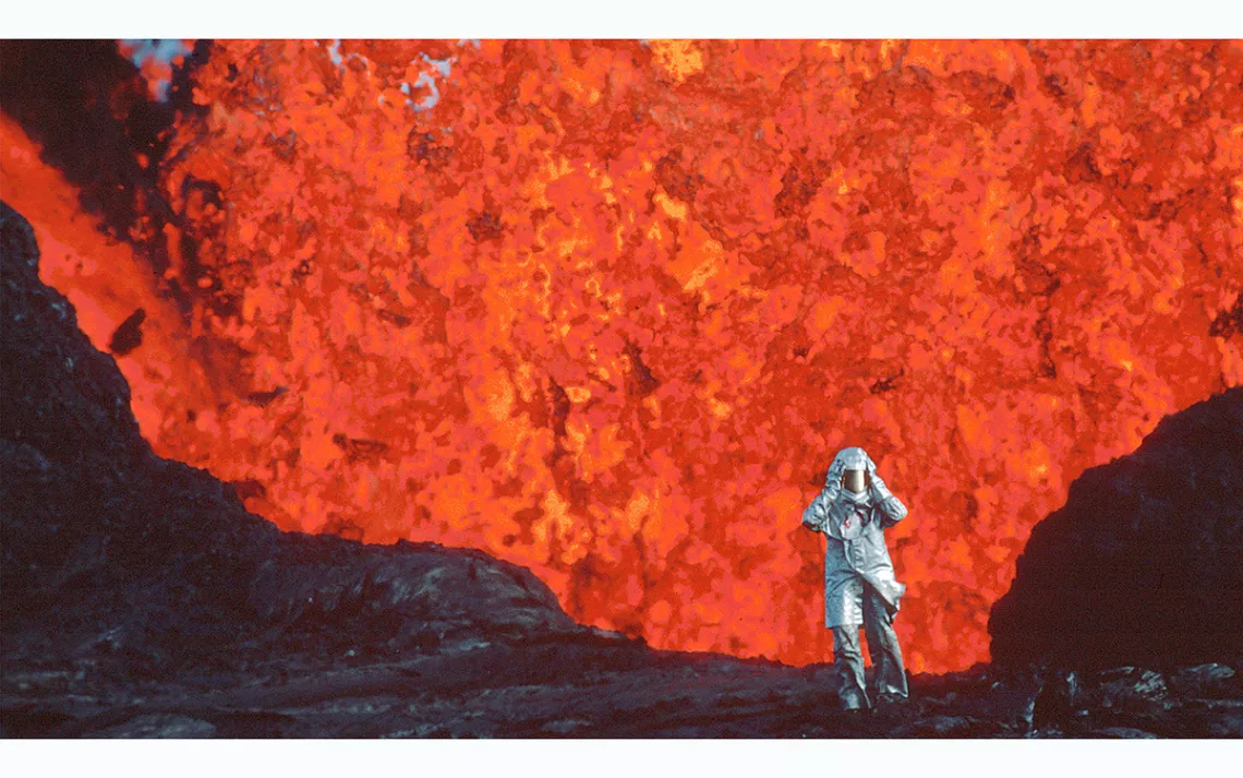A small human figure in a silver foil jumpsuit and helmet stands in front of a fountain of hot red lava.