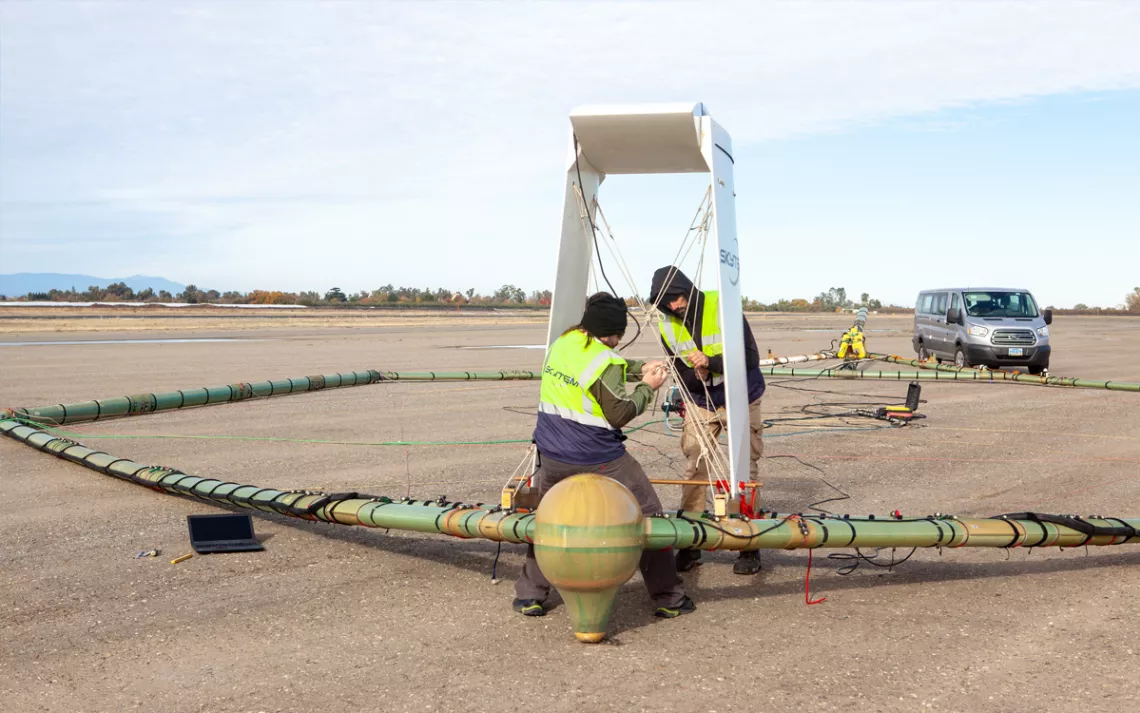Workers in reflective gear make adjustments to a large geometric-shaped piece of equipment on an airfield. 