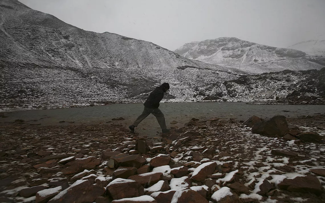 A silhouetted figure walks along a rocky, landscape thinly covered with snow, with white mountains in the distance. 