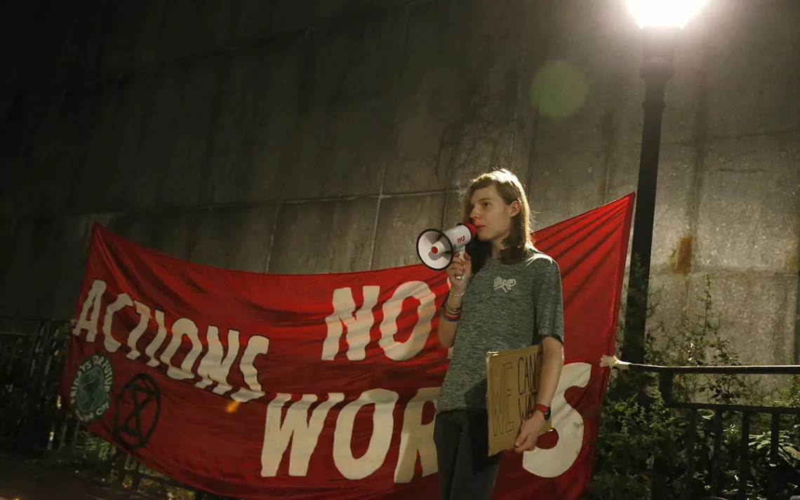 Young woman speaking into a megaphone in front of a red banner that reads "Actions, not Words." 