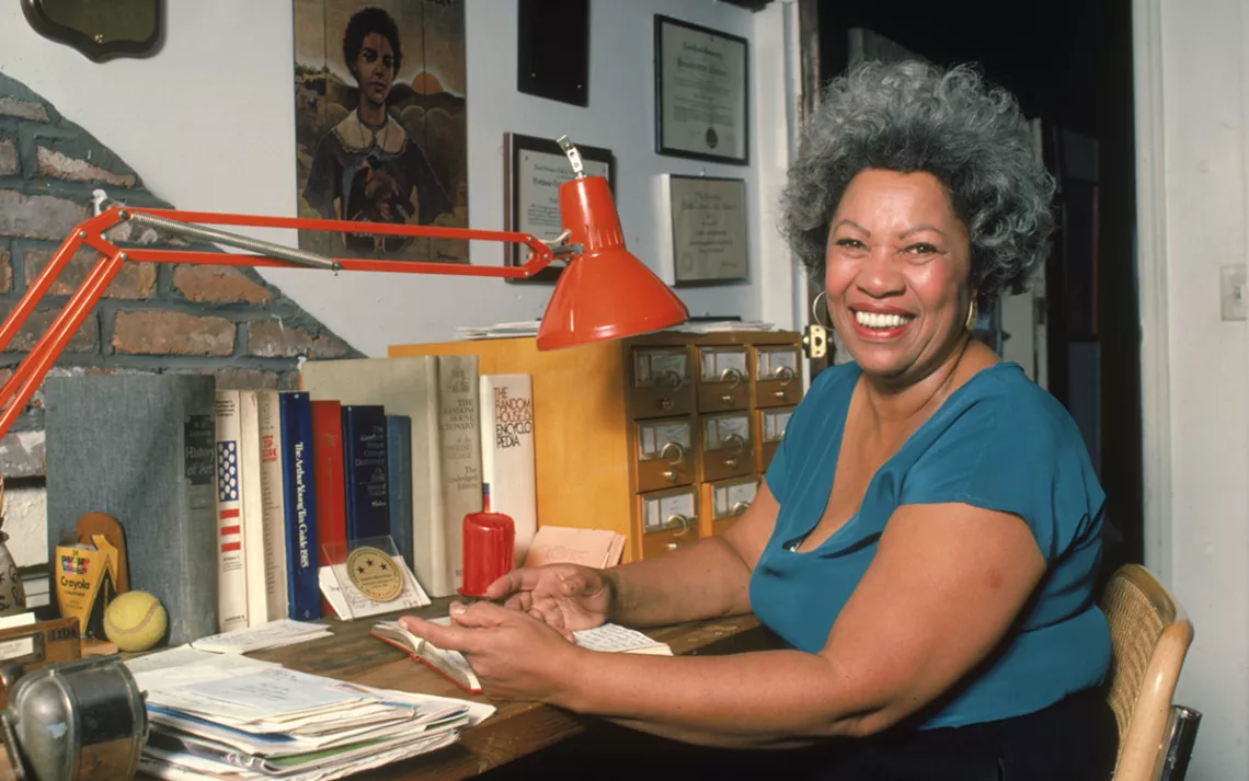 Portrait of American author Toni Morrison smiling and sitting at her desk at home, 1980s.