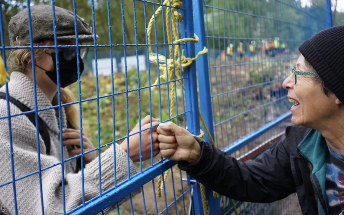Two people—one masked, one not—bump fists through a blue chain-link fence in a forest. 