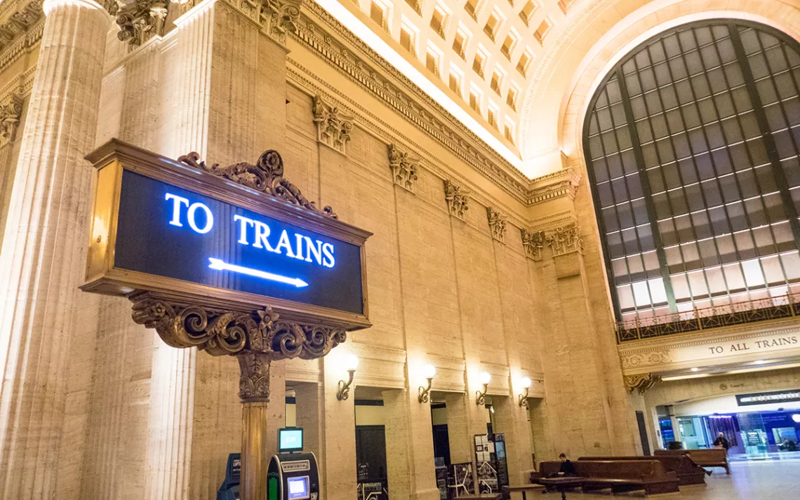 Massive cream-colored stone room in Chicago's Union Station, with an illuminated sign of a blue arrow on a black background reading "to trains." 